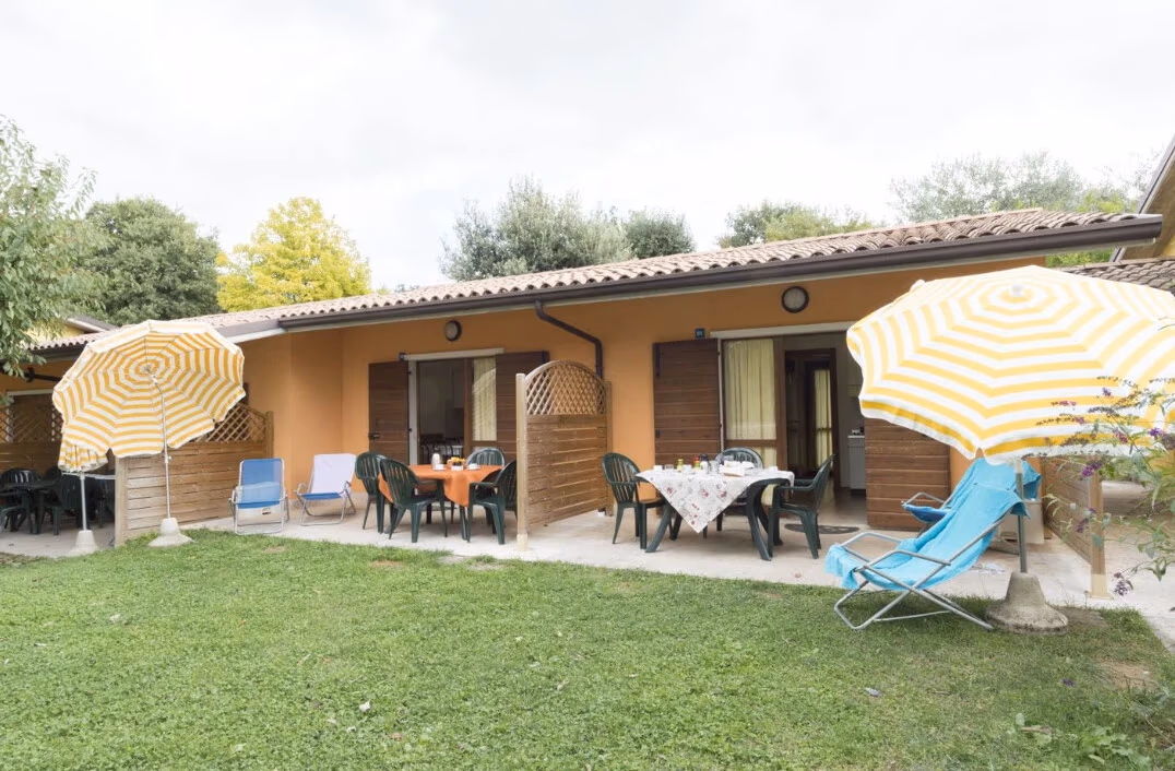 Rij okerkleurige appartementen met pannendak op Camping Piani di Clodia, met geel-wit gestreepte parasols en zitjes in het gras