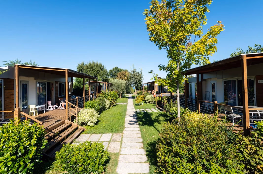 Twee rijen maxicaravans met houten veranda's langs een tegelpad met gras en struiken op Camping Piani di Clodia