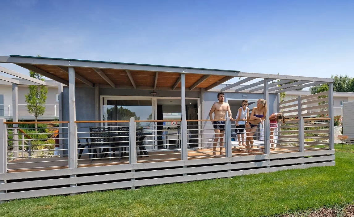Maxicaravan met een grijze gevel en een ruim overdekt terras met eettafel op Camping Piani di Clodia, met gasten op de veranda