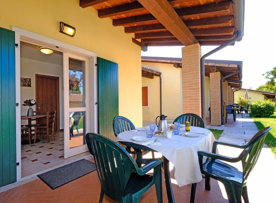 Overdekt terras met een gedekte tafel en groene stoelen bij een geel appartement met groene luiken op Camping Piani di Clodia