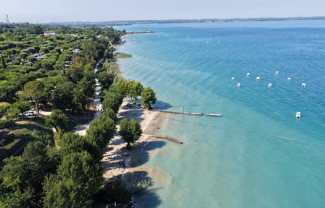 Luchtfoto van de groene oever van Camping Piani di Clodia met strandjes en steigers aan het turquoise Gardameer