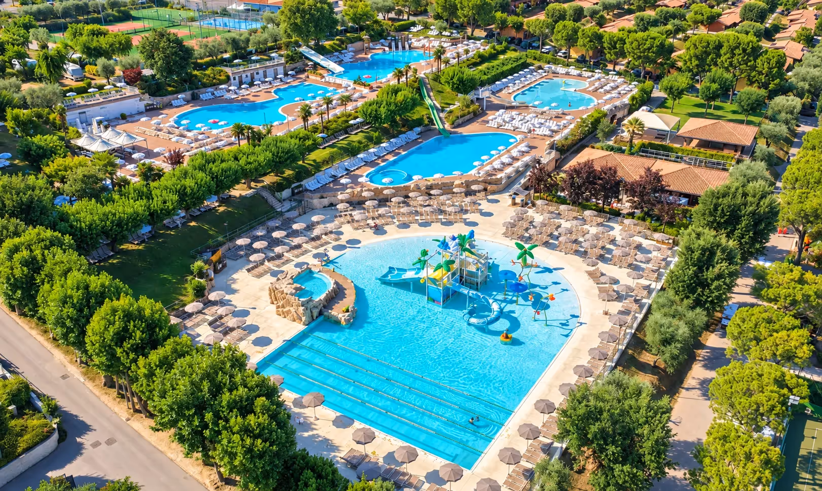 Luchtfoto van de twee waterparken van Camping Piani di Clodia: het wedstrijdbad met banen, het ondiepe kinderbad met een speeltoren in palmboomvorm, de glijbanen en rijen ligbedden met parasols tussen de bomen